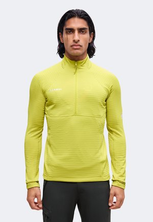 Hombre con cabello oscuro lleva puesto un top de manga larga con cremallera de color verde lima y pantalones negros, de pie de frente contra un fondo claro y sencillo.