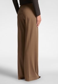Persona che indossa pantaloni marroni a vita alta e larghi, insieme a un top lungo scuro, in piedi con una mano rilassata al fianco.