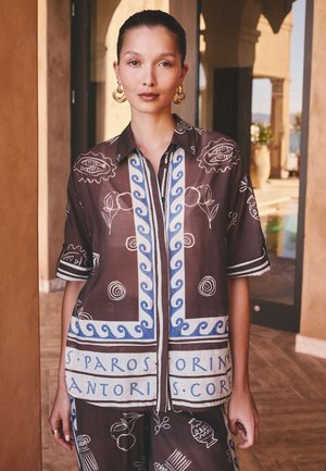 Donna con i capelli tirati indietro indossa una camicia marrone e blu a fantasia e pantaloni abbinati, in piedi in un corridoio ad archi illuminato dal sole.