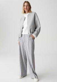 Frau in einer hellgrauen Zip-Jacke, weißem Pullover, weiten grauen Hosen und weißen Sneakers, die mit einer Hand in der Tasche steht.