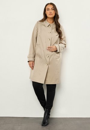 Mujer con abrigo trench beige abotonado, pantalones negros y botines negros, de pie contra una pared blanca lisa.