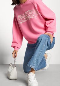 Rosa sweatshirt med "Hollister California" grafik, mysigt tyg och avslappnad passform; kombinerad med ljusblå denim och vita sneakers.