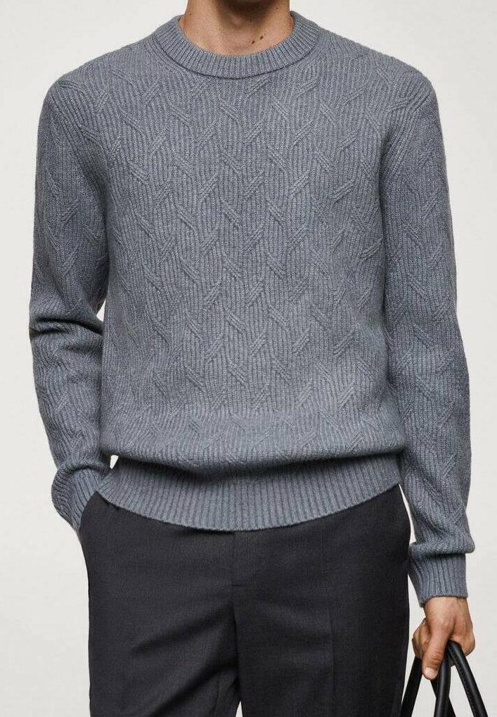 Homme portant un pull en maille grise texturée et un pantalon foncé, tenant un sac noir dans sa main droite, sur un fond uni.