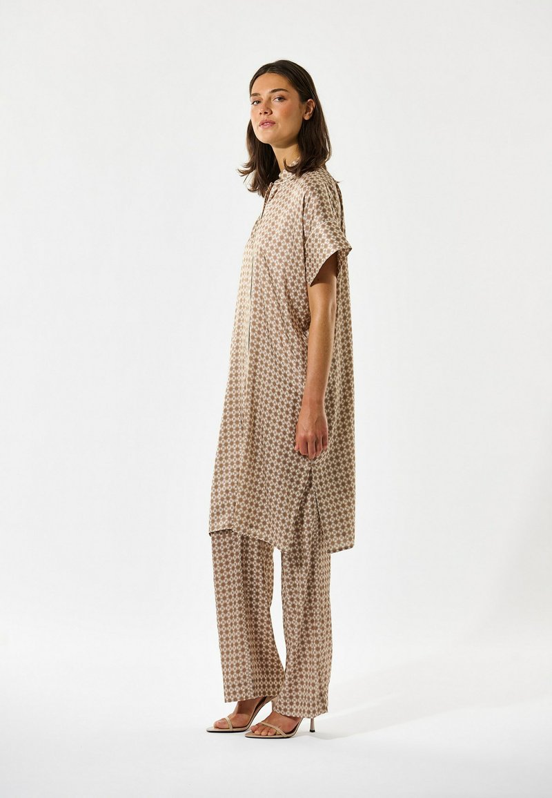 Femme debout, portant une tunique ample beige à motifs avec fentes sur les côtés, un pantalon ample assorti à jambes larges, et des sandales à talons beiges à lanières.