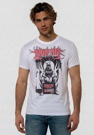 Junger Mann trägt weißes T-Shirt mit schwarzer und roter Grafik einer sitzenden, vermummten Figur, Text: "Boxeur des Rues Boom Bap Beat Starter."