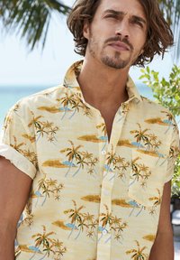 Camicia gialla a maniche corte con bottoni, caratterizzata da una stampa tropicale di palme e isole in verde, marrone e blu su tessuto testurizzato.