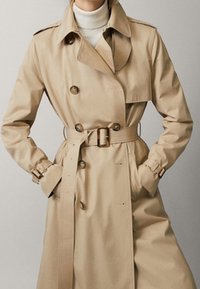Beżowy płaszcz typu trench wykonany z teksturowanej tkaniny, z paskiem w talii, podwójnymi guzikami z przodu i zapięciami na guziki przy mankietach. Klasyczny krój kołnierza.