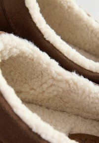 Bruna tofflor med mjukt ullfoder i krämfärg, som visar en slät utsida och en texturerad inre yta för komfort.