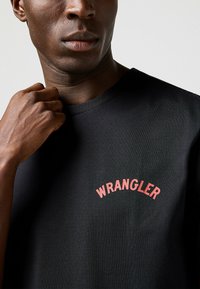 Camiseta de algodón negra con un logo curvado rojo de "WRANGLER" en el pecho. Textura suave con un cuello redondo clásico.