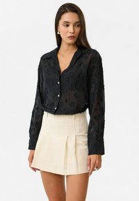 Vrouw in een doorschijnende zwarte blouse met knopen en textuurpatronen, gecombineerd met een hoge taille, geplooide beige minirok.