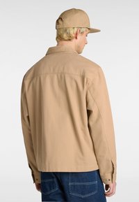 Beige cotton jacket with a pointed collar, back yoke, and button cuffs. Paired with blue denim pants, showing visible stitching and pockets.