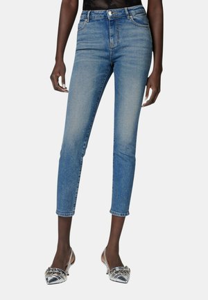 Kvinde iført højtaljede, smalle blå cropped jeans og sølvfarvede spidse slingback-højhælede sko, stående mod en ensfarvet baggrund.