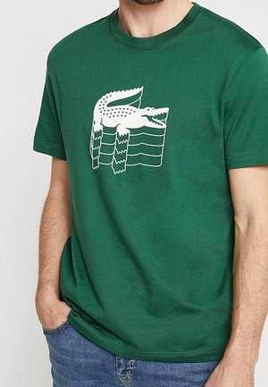 Homme portant un t-shirt vert avec un graphique abstrait de crocodile blanc, assorti à un jean bleu, main dans la poche.