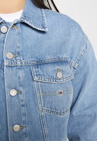 Giubbotto in denim azzurro chiaro con colletto classico, bottoni argentati, due tasche sul petto e una piccola patch con logo in rosso, bianco e blu.