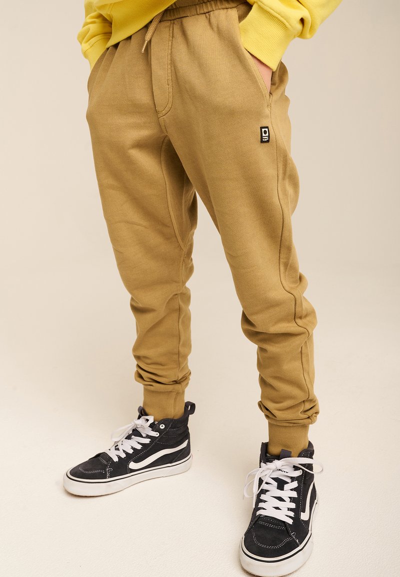 Brune joggerbukser med elastisk talje og manchetter, der har sidelommer og et lille logo-label. Bærer sorte højtop sneakers.