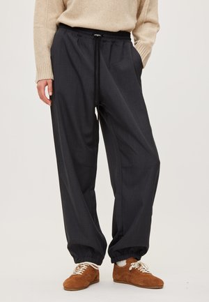 Personne portant un pantalon noir ample avec cordon de serrage et poignets boutonnés, associé à des baskets marron à lacets et un pull beige.