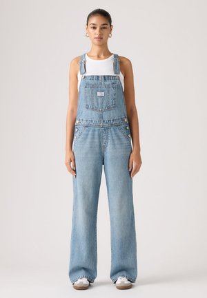 Frau steht und trägt hellblaue Jeans-Latzhose, weißes ärmelloses Oberteil, weiße Turnschuhe und goldene Creolen vor einem einfarbigen Hintergrund.