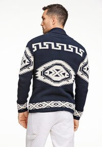 Marinblå stickad cardigan med vita geometriska mönster, vikbar krage och ribbad fåll och ärmslut, designad för en avslappnad passform.