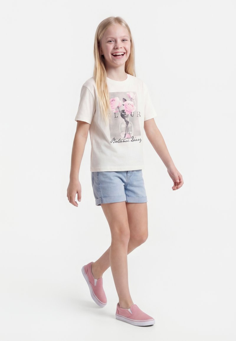 Κορίτσι που χαμογελά περπατά, φορώντας λευκό μπλουζάκι με floral σχέδιο, ανοιχτό μπλε σορτς και ροζ παπούτσια slip-on σε απλό φόντο.