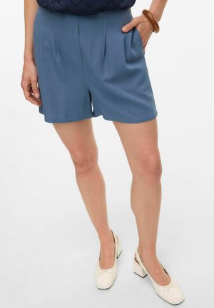 Blauwe op maat gemaakte shorts met plooien, halverwege de bovenbenen, gemaakt van lichtgewicht stof, gecombineerd met witte slingback hakken met strikaccenten.
