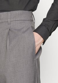 Grijze geribbelde broek met een gladde structuur, gecombineerd met een strak zittend zwart overhemd met patroon. Hand in zak geplaatst, waarbij de taillebanddetails zichtbaar zijn.