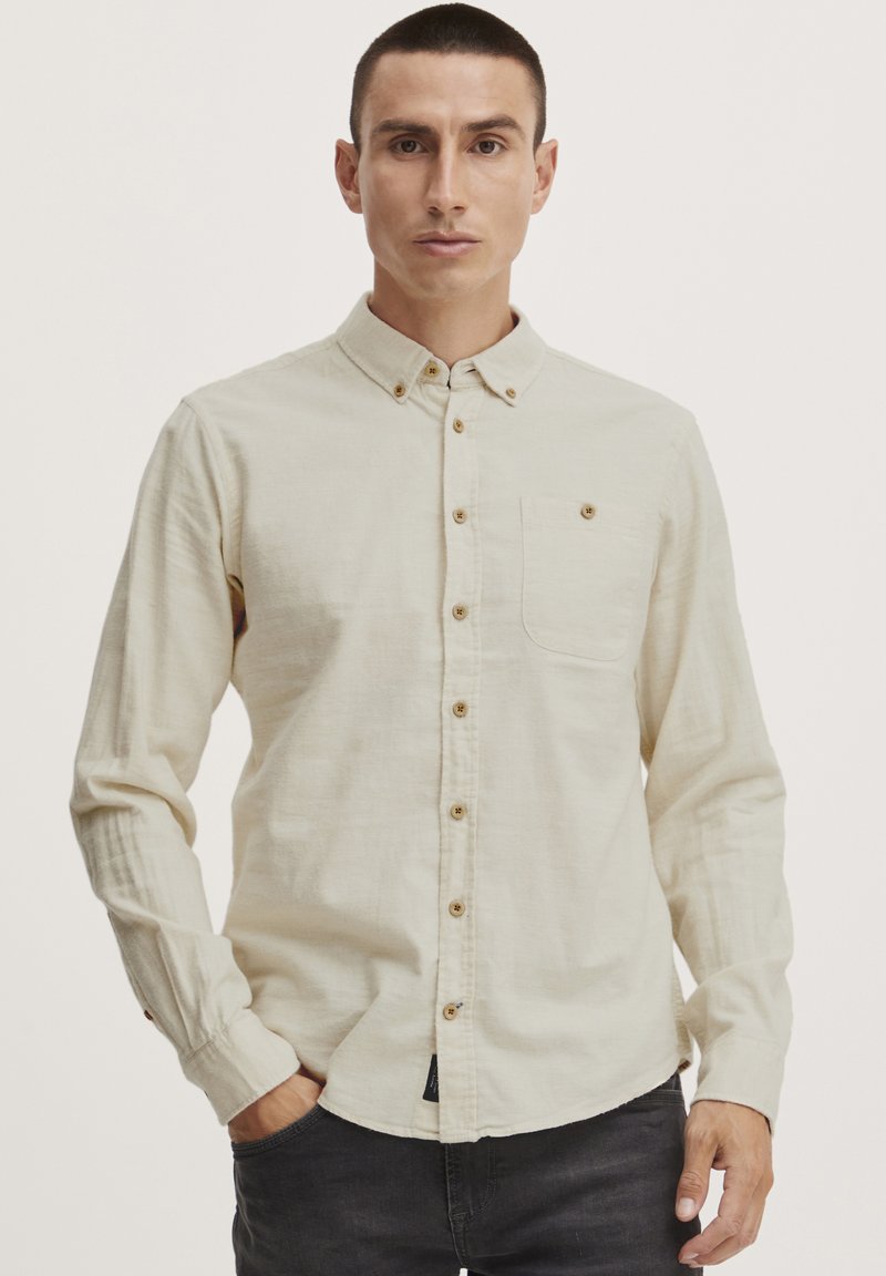 Chemise boutonnée couleur beige clair, avec un col, des manches longues et une poche poitrine. Présente des boutons en bois et un tissu texturé.
