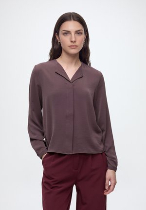 Frau mit langen dunklen Haaren, die eine mauvefarbene langärmelige Bluse und bordeauxrote Hose trägt, steht vor einem schlichten weißen Hintergrund.