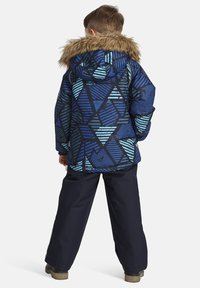 Blått mönstrat pufferjacka med geometriska mönster och fuskpälsfodrad huva; kombinerat med mörkblå snöbyxor. Energisk vinterklädsel.