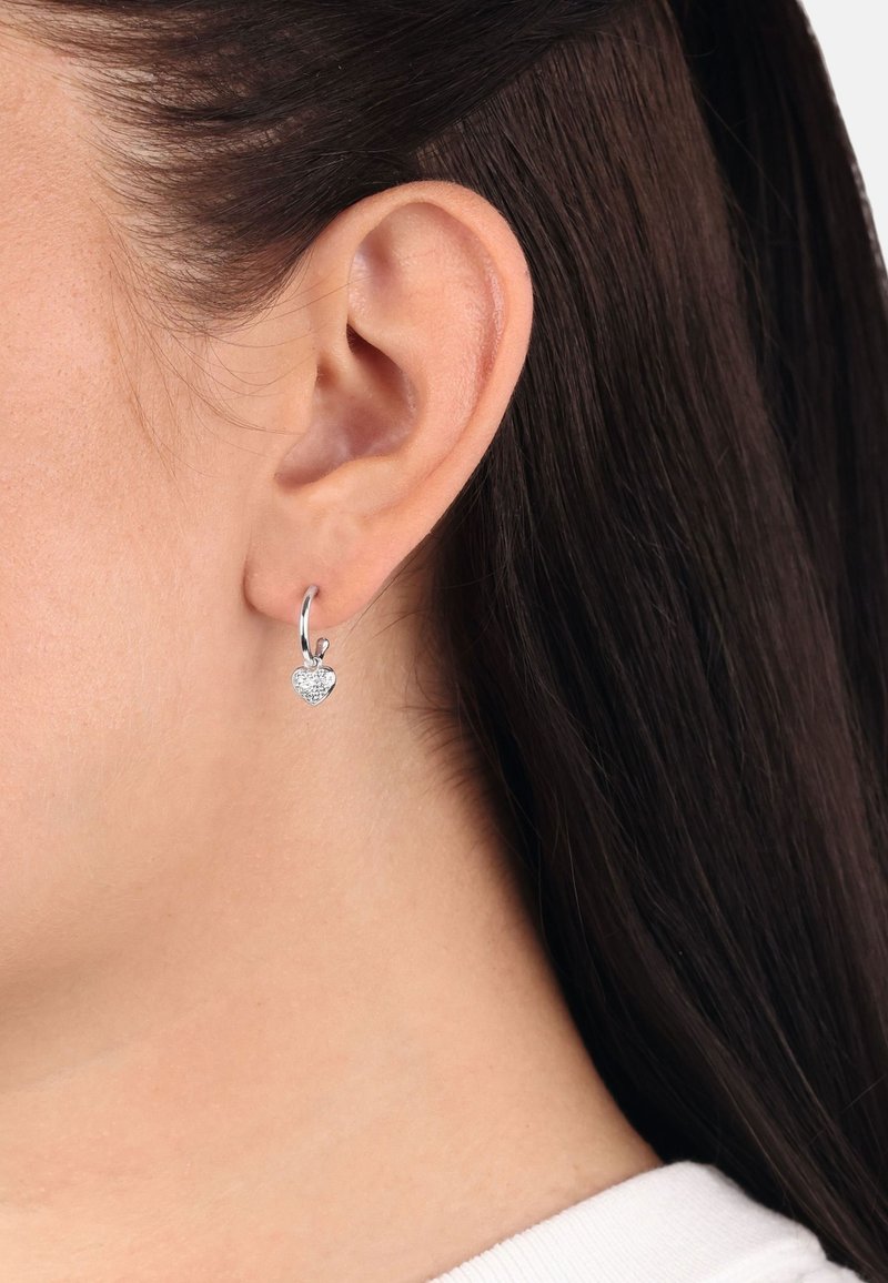 Arete de aro de plata con un colgante en forma de corazón adornado con pequeñas piedras brillantes, mostrado en un modelo con cabello lacio y oscuro.