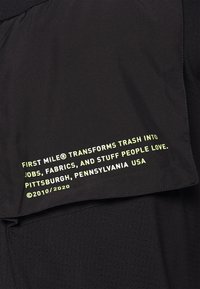 Svart tyg med en texturerad yta som har en framträdande grön tryckt etikett som står: "FIRST MILE® förvandlar skräp till jobb..."