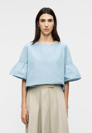 Γυναίκα με ίσια σκούρα μαλλιά φοράει ανοιχτό μπλε μπλούζα με φαρδιά μανίκια με βολάν και μπεζ πλισέ παντελόνι ψηλόμεσο, μπροστά σε απλό φόντο.