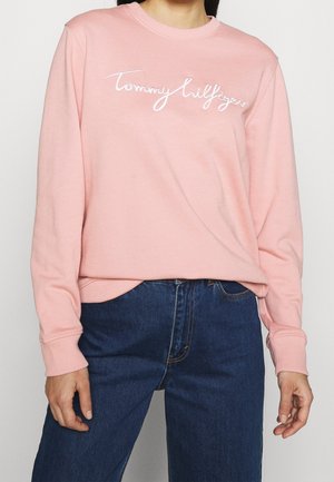 Person iført en lyserød langærmet sweatshirt med teksten "Tommy Hilfiger" og blå cowboybukser, stående mod en ensfarvet baggrund.