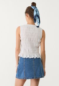 Weißes, gerafftes, ärmelloses Oberteil mit einem Rüschensaum, kombiniert mit einem kurzen blauen Denimrock. Das Model trägt einen gemusterten blauen Schal im Haar.