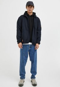 Marinefarget fleecejakke med glidelås, lagd over en svart hettegenser, parret med blå jeans og grå sneakers, komplementert av en mørk caps.