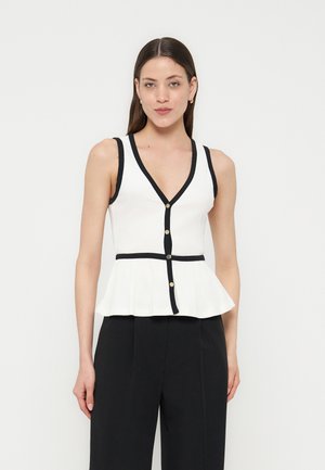 Femme portant un top blanc sans manches avec des finitions et des boutons noirs, associé à un pantalon noir taille haute, se tenant devant un fond uni.