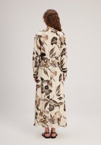 Robe maxi en tissu beige avec de grands motifs floraux marron, à manches longues et taille ceinturée, portée avec des sandales noires plates.