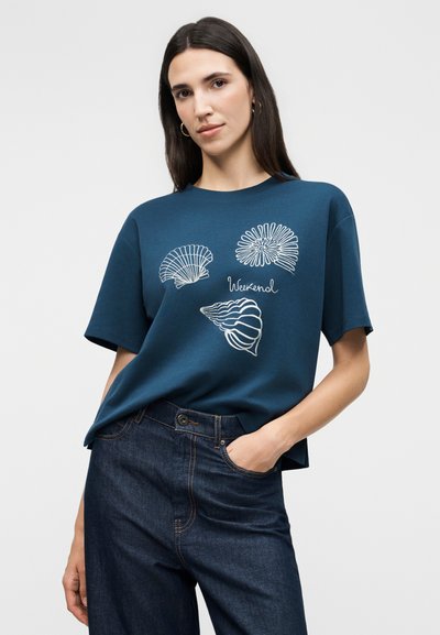 Νεαρή γυναίκα που φοράει μπλε μπλούζα με λευκά σχέδια κοχυλιών και την επιγραφή "Weekend", συνδυασμένη με σκούρο μπλε τζιν, ποζάροντας με το ένα χέρι στην τσέπη.