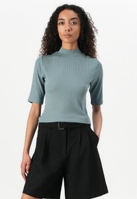 Teal ribbatoppi, jossa on mock neck ja lyhyet hihat, yhdistettynä mustiin pleatattuihin shortseihin, joissa on suorakulmainen solkilöysä.