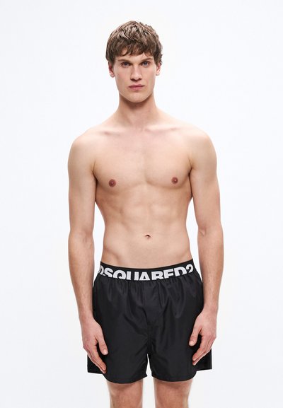 Shorts de bain noirs avec une ceinture élastique blanche portant un logo contrastant. Le tissu semble lisse, léger et à séchage rapide. Le modèle pose de face.