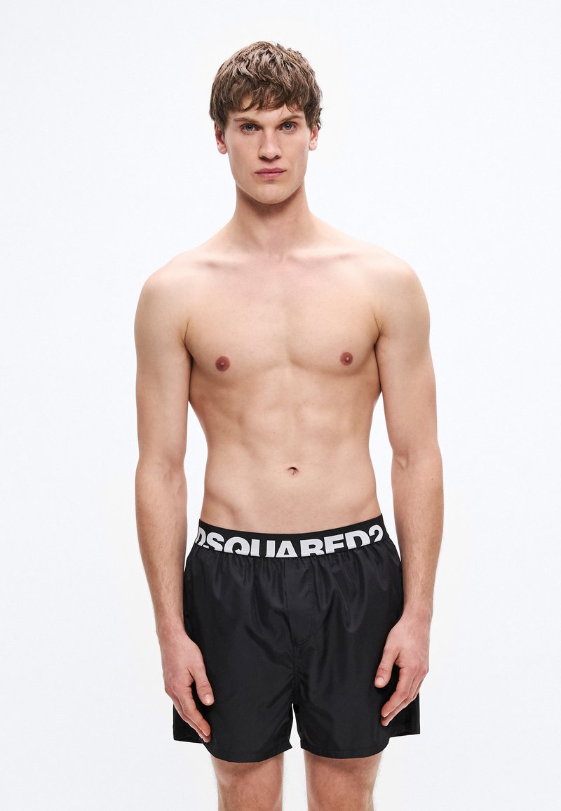 Bañador corto negro con una cintura elástica blanca que lleva un logo en contraste. La tela parece suave, ligera y de secado rápido. El modelo posa de frente.