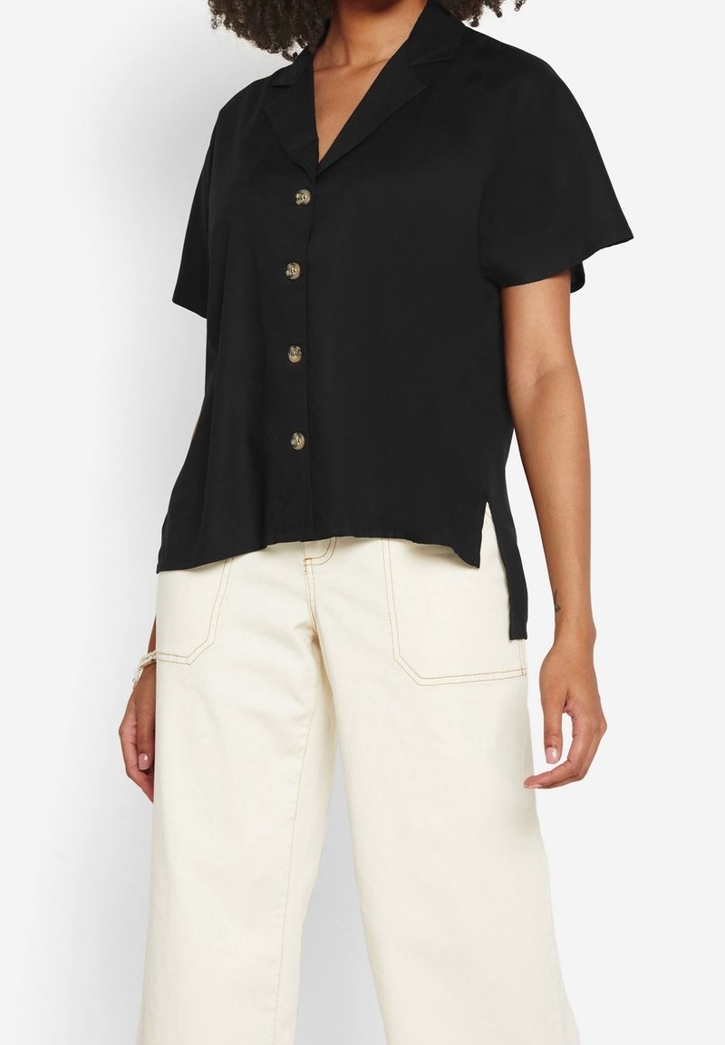 Black short-sleeve shirt with a collar, five brass buttons, and side slits, made from smooth fabric. Paired with cream-colored wide-leg pants.