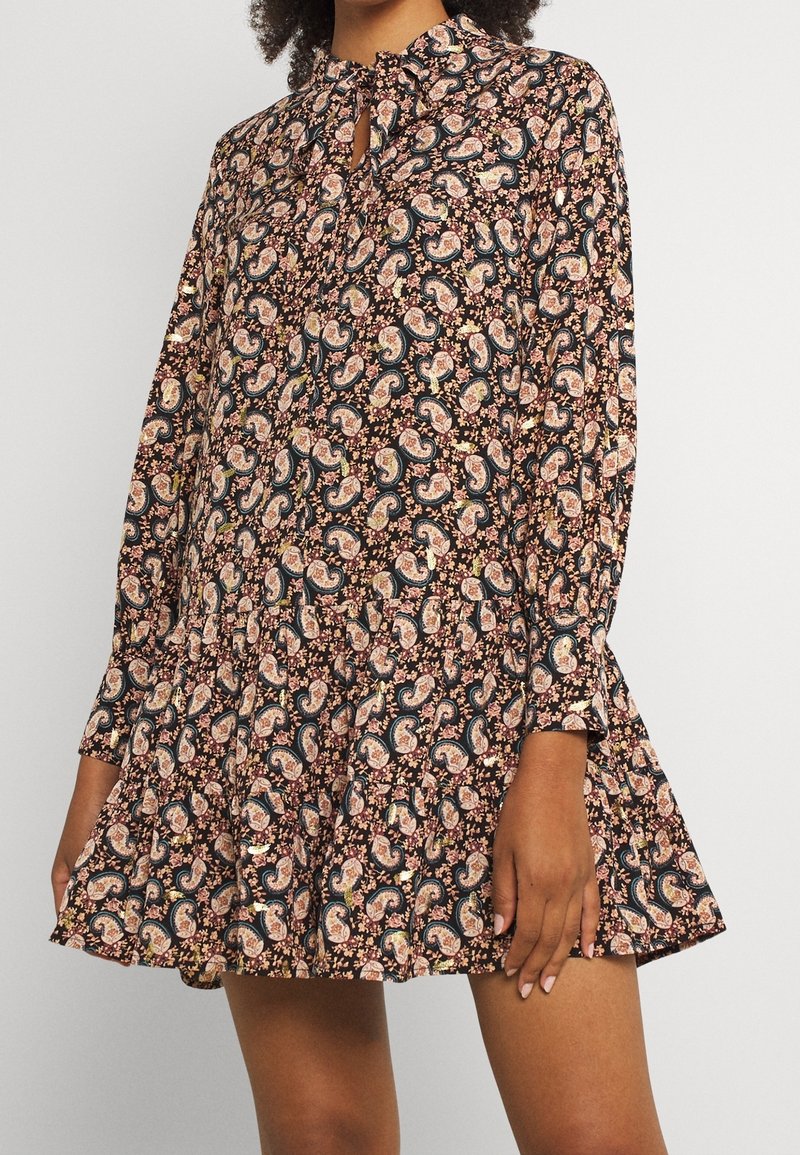 Femme portant une robe courte à manches longues avec un motif cachemire multicolore et floral sur un fond sombre.