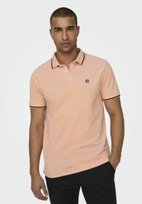 Camisa polo rosa claro con ribetes negros en el cuello y las mangas, con un logotipo en el pecho, hecha de un material de algodón suave.