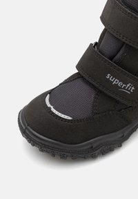 Botte noire pour enfants avec un extérieur texturé, des sangles velcro et une semelle en caoutchouc. Comprend des panneaux en maille et des accents réfléchissants.