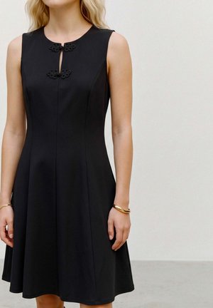 Un mannequin porte une robe noire sans manches à coupe trapèze, ornée de deux fermetures décoratives chinoises et de bracelets dorés.