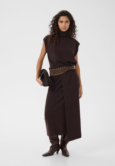 Femme aux cheveux bouclés portant une robe marron foncé sans manches à col roulé, ceinture cloutée, tenant une pochette noire et des bottes à talons marron.