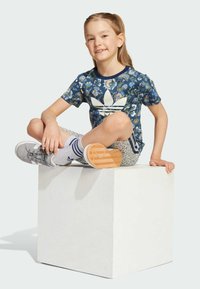 Farebná mládežnícka tričko s kvetinovým vzorom v námorníckej modrej s bielym logom Adidas, spárované s vzorovanými šortkami a sivými teniskami na bielom kubuse.