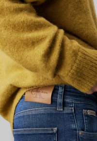 Horčicovožltý sveter s textúrovanou látkou, skombinovaný s tmavomodrými džínsami s viditeľnou koženou nášivkou s nápisom "Calvin Klein Jeans."