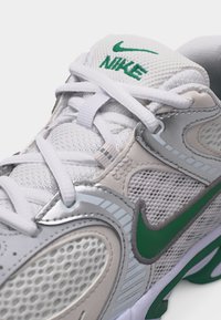 Fehér és szürke Nike sportcipő zöld pipa logókkal, hálós panelek és fehér fűzők, melyek párnázott nyelv fölött, a hímzett márkanévvel vannak megkötve.