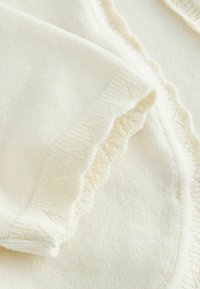 Tissu tricoté doux de couleur crème avec bords repliés présentant des volants délicats et des motifs côtelés texturés.